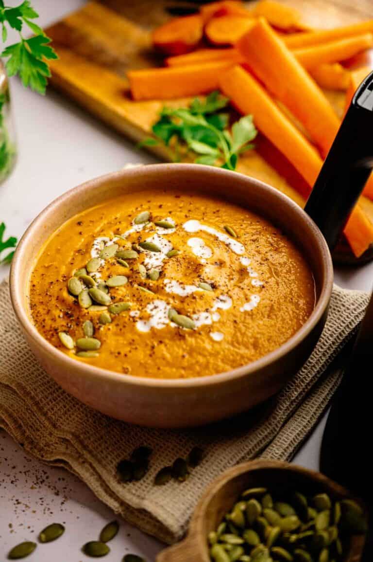 A bowl of creamy Air Fryer Vegan Carrot Soup topped with a swirl of cream and pumpkin seeds, with fresh carrot sticks and herbs in the background.