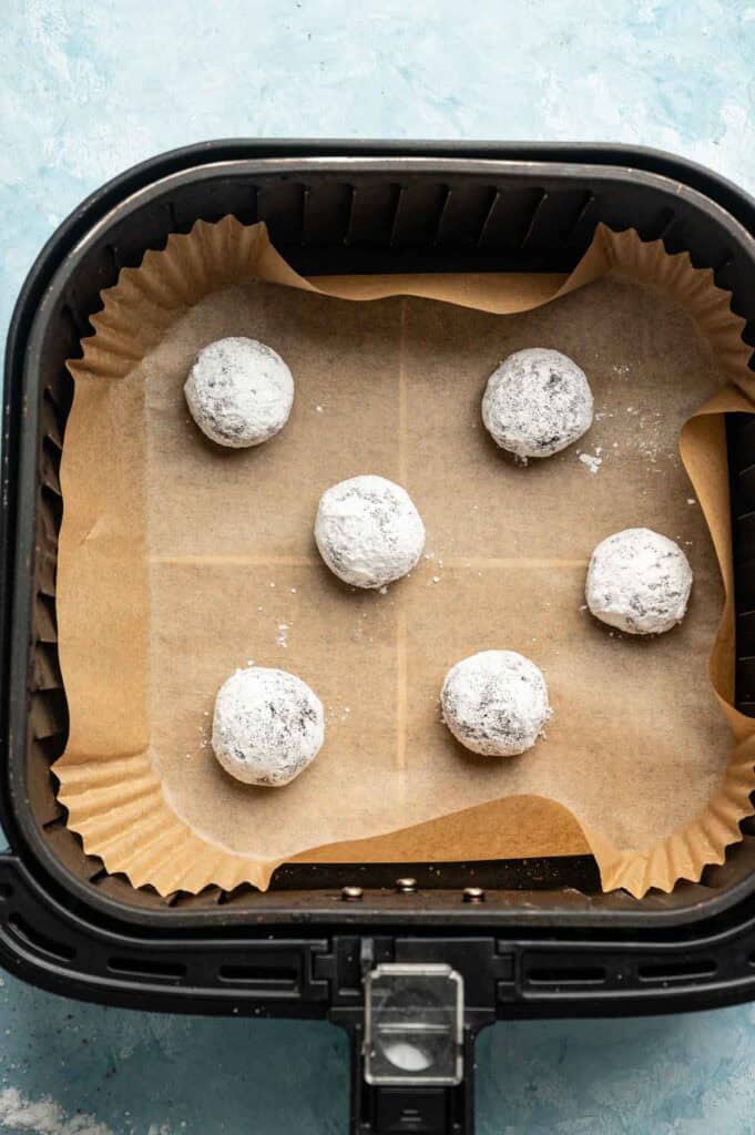 Six Air Fryer Vegan Crinkle Cookies, shaped into powdered dough balls, rest on parchment paper inside the air fryer basket.