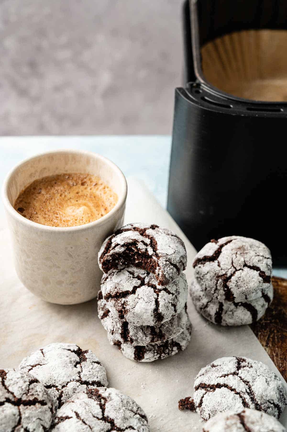 A cup of coffee sits next to a stack of Air Fryer Vegan Crinkle Cookies dusted with powdered sugar, with an air fryer in the background.