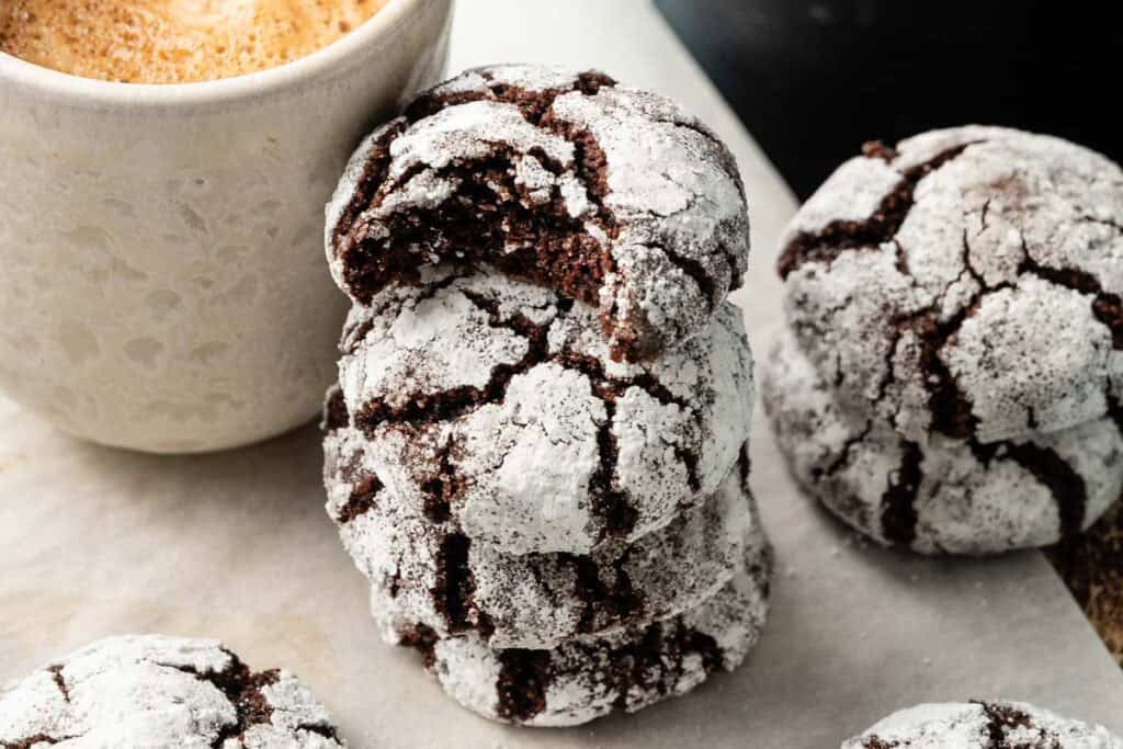 A stack of Air Fryer Vegan Crinkle Cookies dusted with powdered sugar, one with a bite taken out, sits next to a cup of coffee on a light surface.