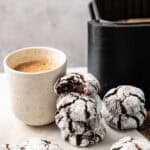 A cup of coffee sits next to a stack of Air Fryer Vegan Crinkle Cookies, with an air fryer in the background and more cookies scattered on the surface.