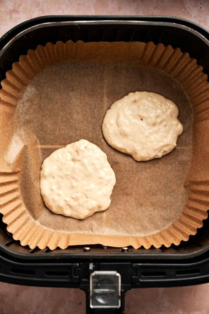 Two dollops of Air Fryer Vegan Pancakes batter sit on parchment paper inside an air fryer basket, ready to be cooked.
