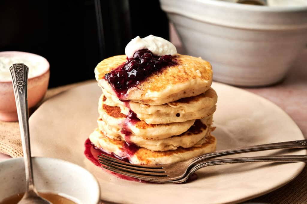 A stack of Air Fryer Vegan Pancakes topped with berry sauce and a dollop of whipped cream sits on a plate with a fork, surrounded by bowls and a cup.