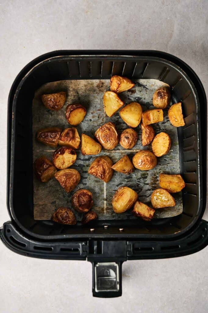 Air Fryer Vegan Roasted Potatoes, cut and perfectly browned, are arranged at the bottom of an air fryer basket lined with parchment paper.