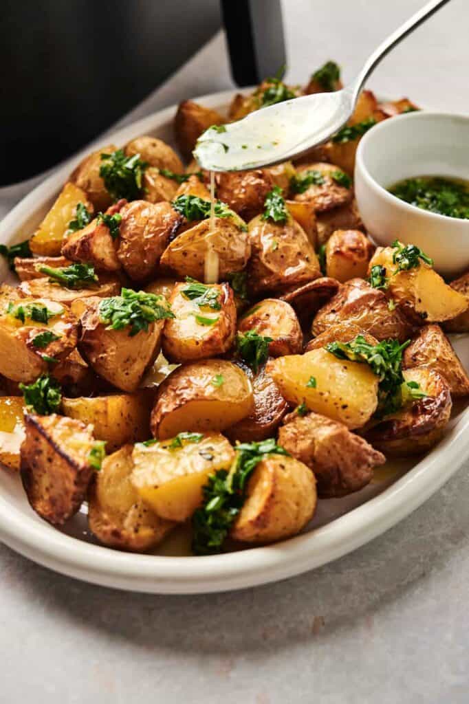 Air Fryer Vegan Roasted Potatoes are served as golden wedges on a white plate, garnished with chopped herbs. A spoon drizzles savory sauce over the potatoes, with a small bowl of vibrant green sauce on the side.