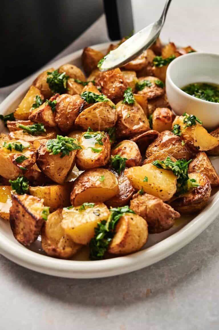 A plate of Air Fryer Vegan Roasted Potatoes topped with chopped herbs, next to a small bowl of green sauce; a spoon hovers above, drizzling sauce.