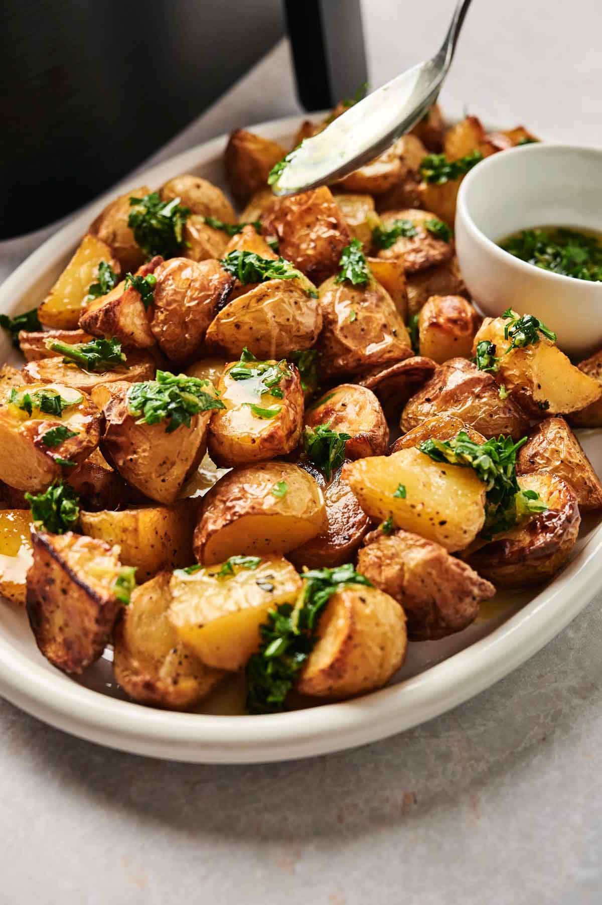 A plate of Air Fryer Vegan Roasted Potatoes topped with chopped herbs, next to a small bowl of green sauce; a spoon hovers above, drizzling sauce.