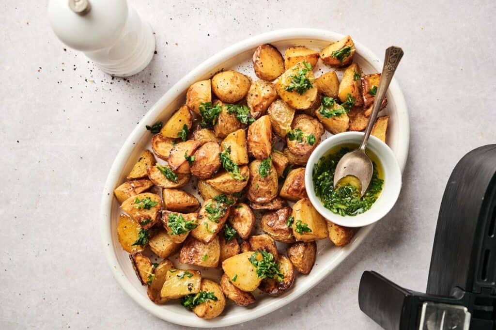Oval platter of Air Fryer Vegan Roasted Potatoes garnished with chopped herbs, served with a bowl of green sauce and a spoon; a pepper grinder is nearby.