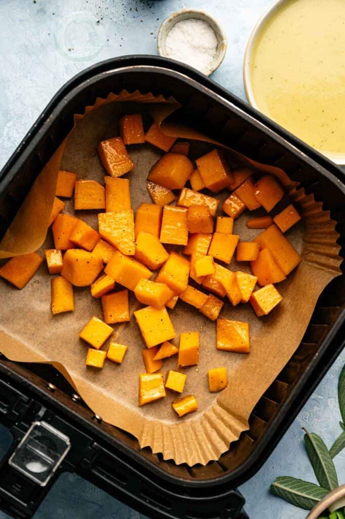 Cubed butternut squash pieces on parchment paper inside an air fryer basket, seasoned and ready to be cooked for a delicious Air Fryer Vegan Squash Casserole.