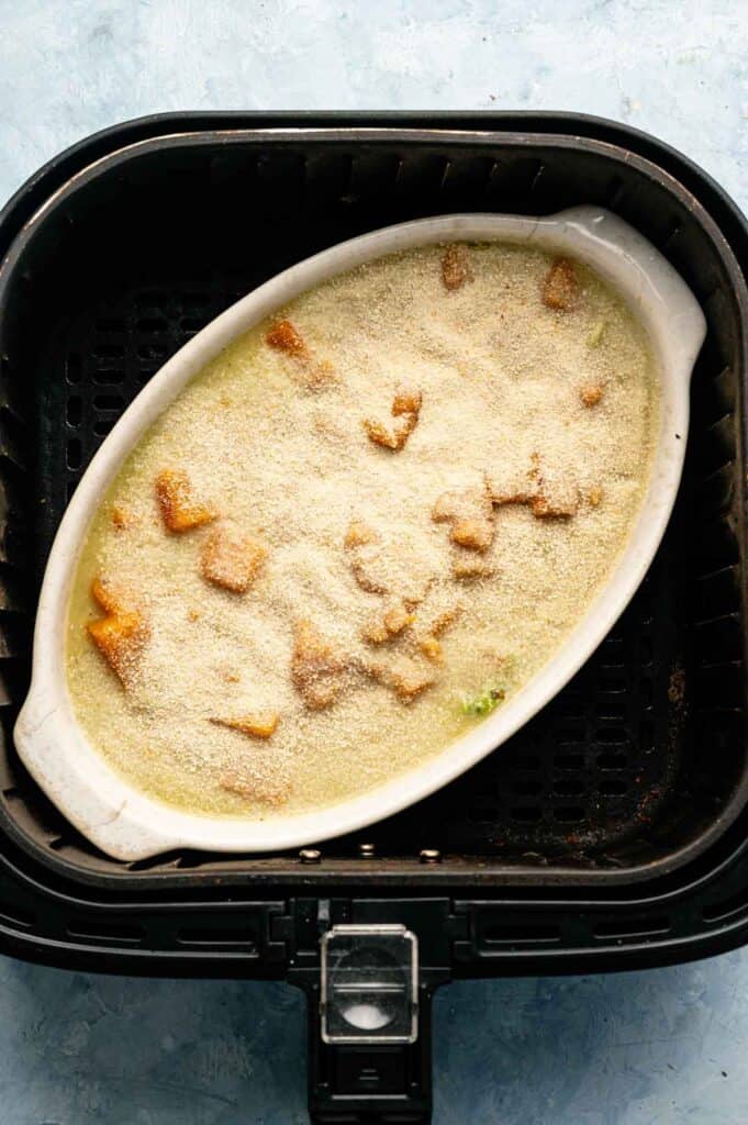 An oval white dish filled with Air Fryer Vegan Squash Casserole, topped with golden breadcrumbs, sits inside an air fryer basket on a light blue surface.