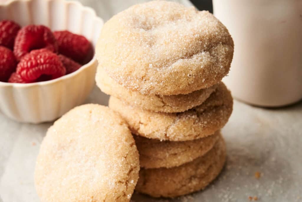 A stack of Air Fryer Vegan Sugar Cookies sits on a surface next to a bowl of raspberries and part of a white cup.