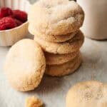 A stack of Air Fryer Vegan Sugar Cookies sits on parchment paper, with a small bowl of raspberries and a white cup in the background. Crumbs are scattered in the foreground.