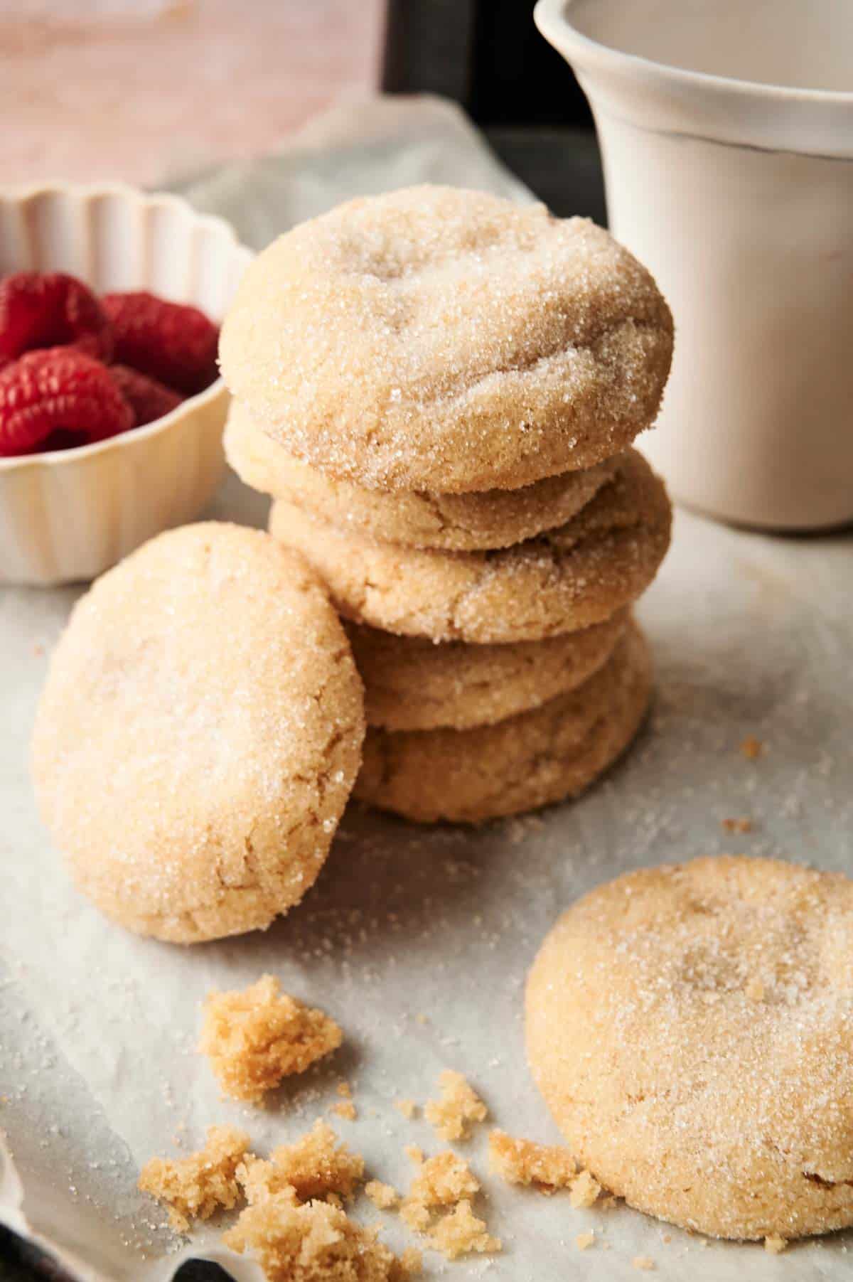 A stack of Air Fryer Vegan Sugar Cookies rests on parchment paper, with a small bowl of raspberries and a white cup in the background. Crumbs are scattered nearby.