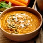 A bowl of creamy Air Fryer Vegan Carrot Soup garnished with a swirl of cream, pumpkin seeds, and cracked black pepper, with vegetables blurred in the background.