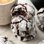 A stack of three Air Fryer Vegan Crinkle Cookies dusted with powdered sugar, with a bite taken from the top cookie, sits beside a light-colored mug.