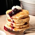 A stack of Air Fryer Vegan Pancakes topped with berry sauce and a dollop of cream sits on a plate with a fork.