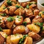 Air Fryer Vegan Roasted Potatoes wedges topped with chopped herbs and a drizzle of sauce, served on a white plate with a small bowl of green sauce on the side.