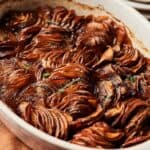 A baked dish of thinly sliced potatoes arranged in overlapping layers, browned and garnished with fresh herbs in a white oval baking dish, reminiscent of a Sweet Potato Casserole with Orange Juice.