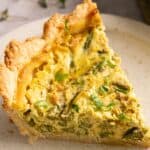 A slice of vegan asparagus quiche with a flaky crust, topped with chopped herbs, served on a beige plate.
