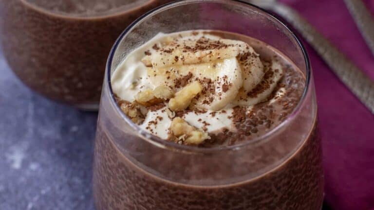 A glass of chocolate chia pudding topped with banana slices, chopped walnuts, a dollop of cream, and chocolate shavings.