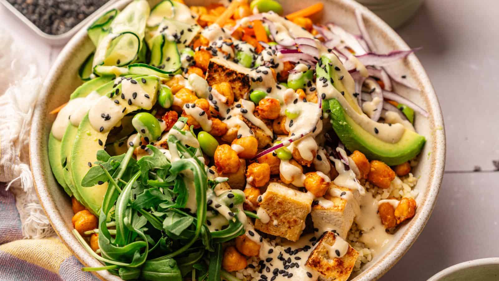 A bowl filled with tofu, chickpeas, avocado slices, arugula, edamame, shredded vegetables, and black sesame seeds, topped with a creamy dressing.