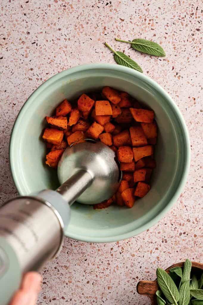 An immersion blender blending roasted pumpkin in a light green bowl on a speckled countertop, with fresh sage leaves nearby.