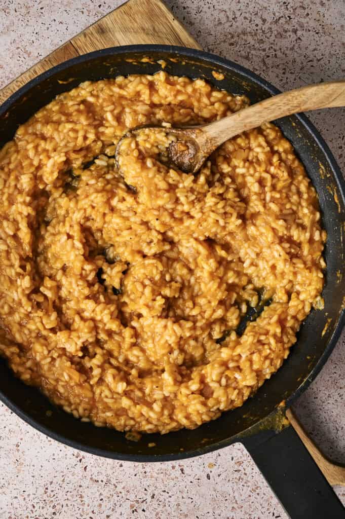 A pan filled with risotto rice coated in smooth pumpkin puree is being stirred with a wooden spoon on a speckled countertop.