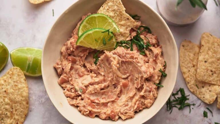 A bowl of refried beans garnished with lime slices and cilantro, with tortilla chips placed around and inside the bowl on a light surface.