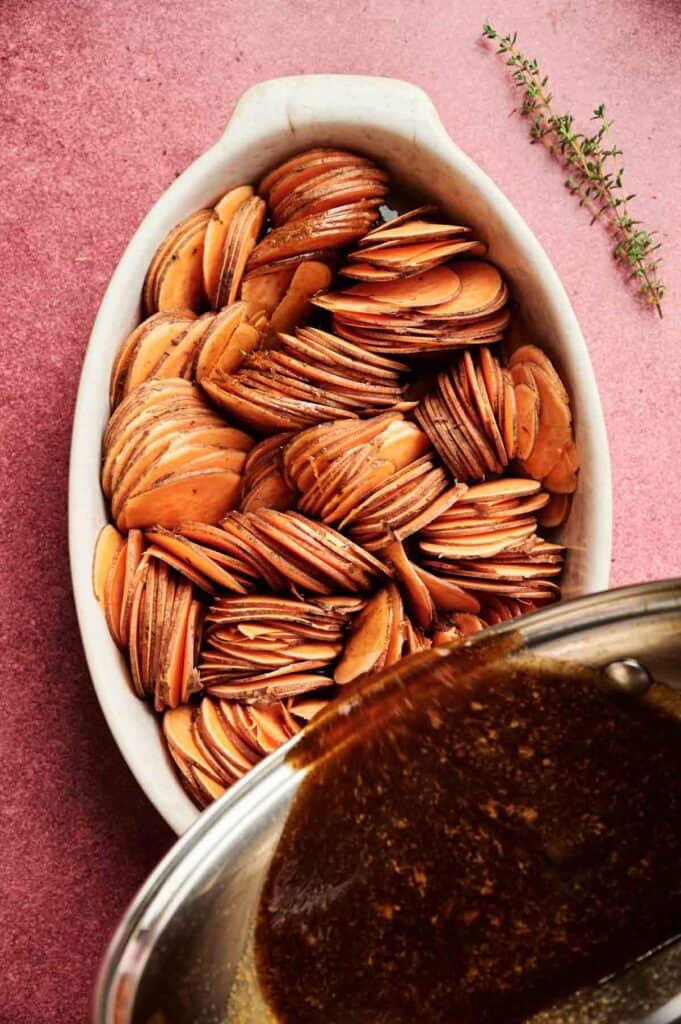 Thinly sliced sweet potatoes are arranged upright in a white oval baking dish, with a luscious sauce—highlighting the zesty brightness of orange juice—being poured over them; thyme sprigs rest beside the dish, capturing the inviting essence of this Sweet Potato Casserole with Orange Juice.