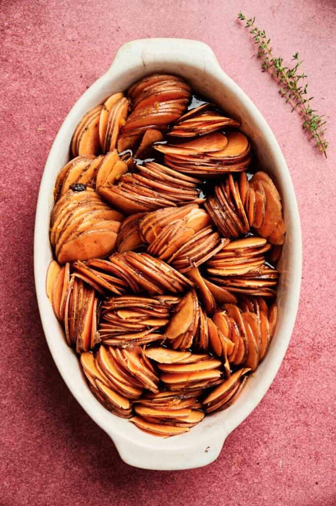 Sliced potatoes arranged in a white oval baking dish with sprigs of thyme on a pinkish surface, reminiscent of a Sweet Potato Casserole with Orange Juice.