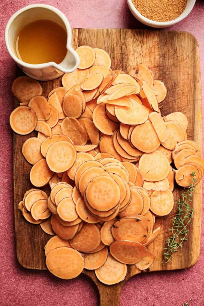 Thinly sliced sweet potatoes are arranged on a wooden cutting board with a sprig of thyme, a small pitcher of oil, and a bowl of seasoning nearby—perfect ingredients for preparing a flavorful Sweet Potato Casserole with Orange Juice.