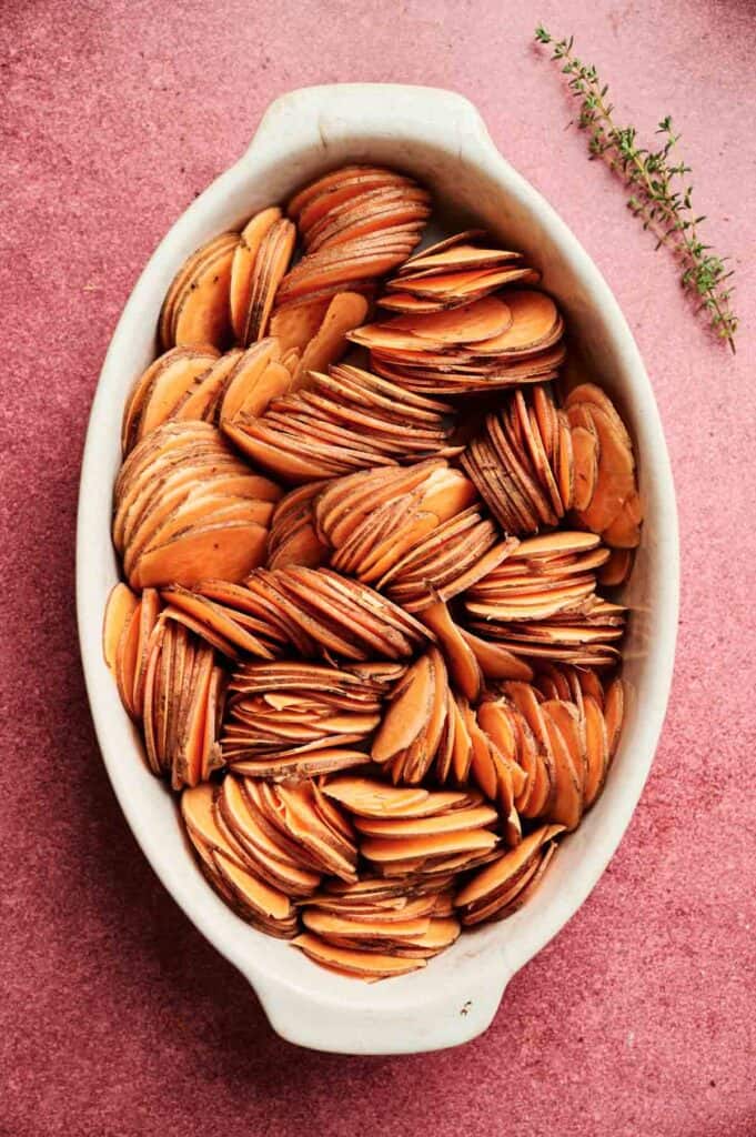 Thinly sliced sweet potatoes arranged vertically in a white oval baking dish, with a sprig of fresh thyme next to the dish on a pink surface—this inviting Sweet Potato Casserole with Orange Juice promises vibrant flavor and elegant presentation.