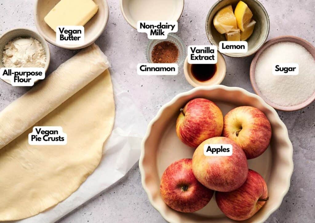 A flat lay of labeled ingredients for Air Fryer Vegan Apple Pie, including pie crusts, apples, sugar, lemon, vanilla extract, cinnamon, non-dairy milk, vegan butter, and flour.
