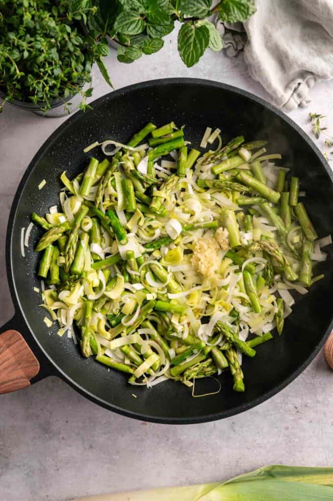 Sliced asparagus, leeks, and minced garlic cooking in a black skillet on a light surface, with fresh herbs visible nearby—perfect savory ingredients for a Vegan Asparagus Quiche.