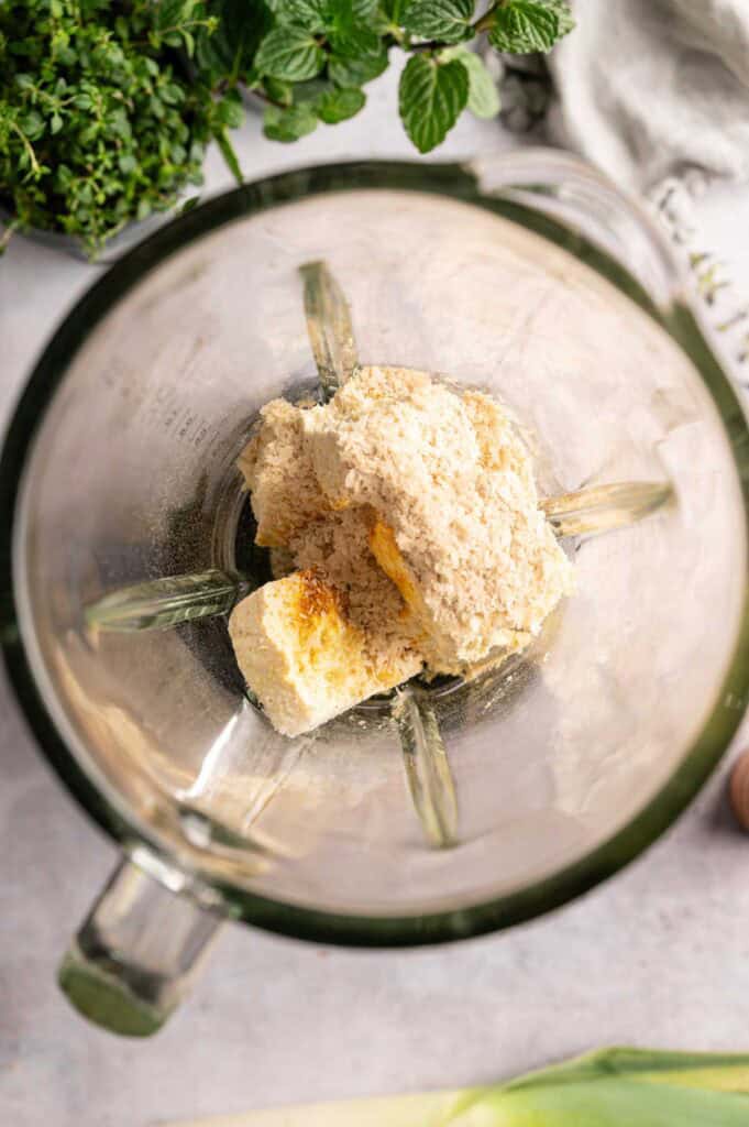 A blender with scoops of vanilla ice cream inside, viewed from above, sits on the counter with fresh herbs and a cloth in the background—perfect inspiration for experimenting with flavors like those found in a Vegan Asparagus Quiche.