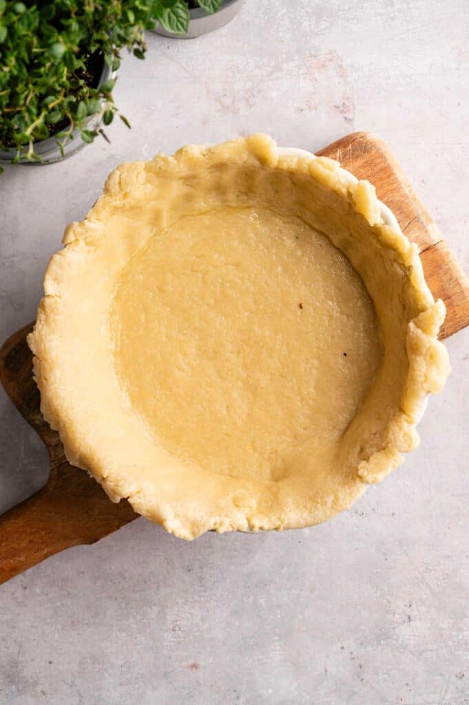 Unbaked pie crust in a round dish on a wooden board, perfect as the base for a Vegan Asparagus Quiche, with herbs in the top left corner on a light surface.