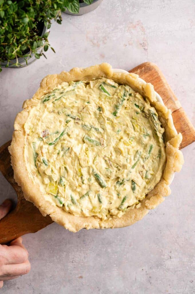 Vegan Asparagus Quiche with an unbaked vegetable filling and a rustic pie crust in a white dish, placed on a wooden board next to potted herbs on a light surface.