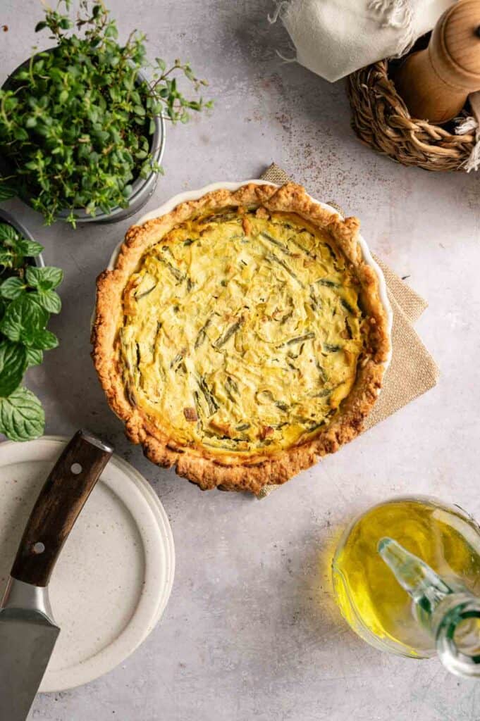A Vegan Asparagus Quiche in a pie dish on a light surface, surrounded by fresh herbs, a knife on a plate, and a bottle of olive oil.