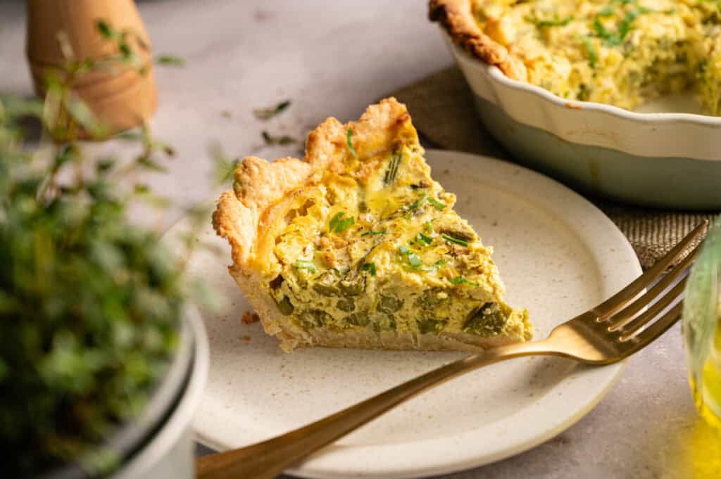 A slice of Vegan Asparagus Quiche on a white plate with a fork, with a potted herb and the rest of the quiche in the background.