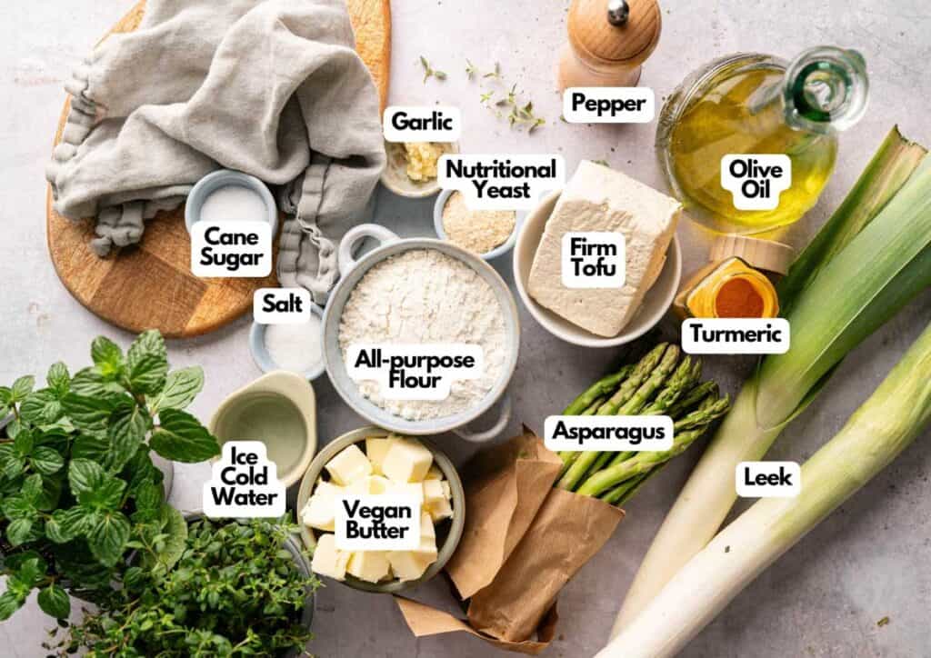 Top-down view of labeled ingredients for an Air Fryer Vegan Asparagus Quiche, featuring flour, tofu, vegan butter, olive oil, leek, asparagus, nutritional yeast, spices, herbs, sugar, and water.