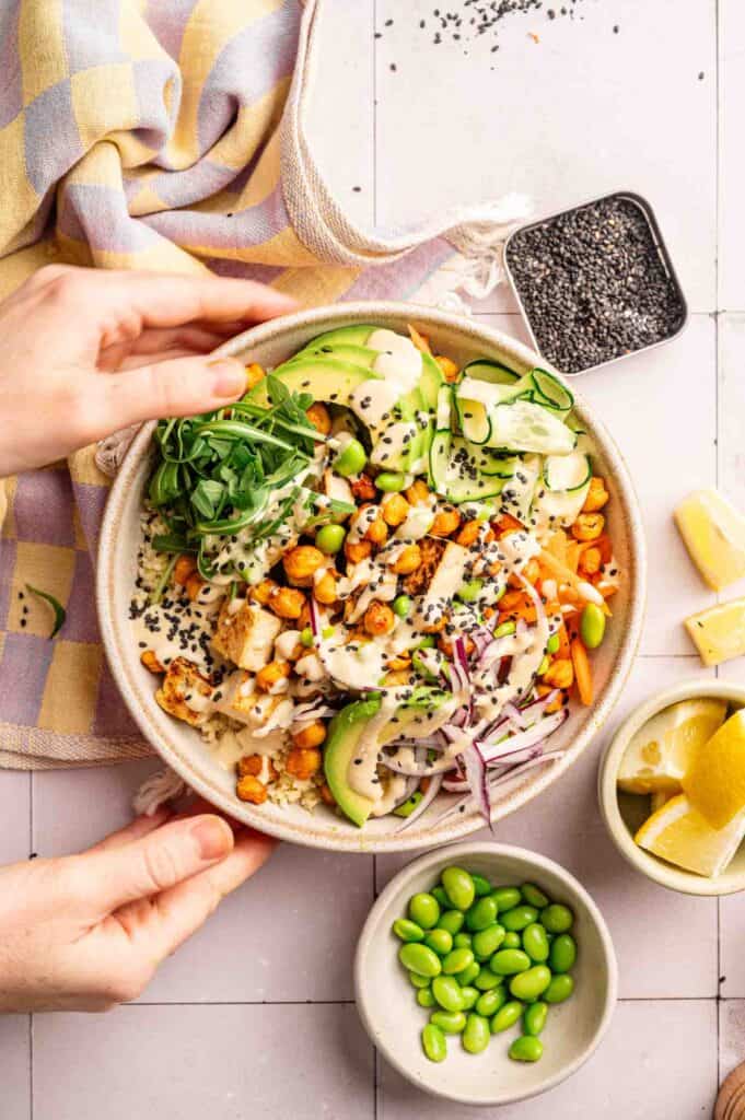 A person holds a vibrant Vegan Buddha Bowl filled with vegetables, chickpeas, avocado, greens, sesame seeds, and creamy dressing. Lemon wedges and edamame are in small bowls nearby on a tiled surface.