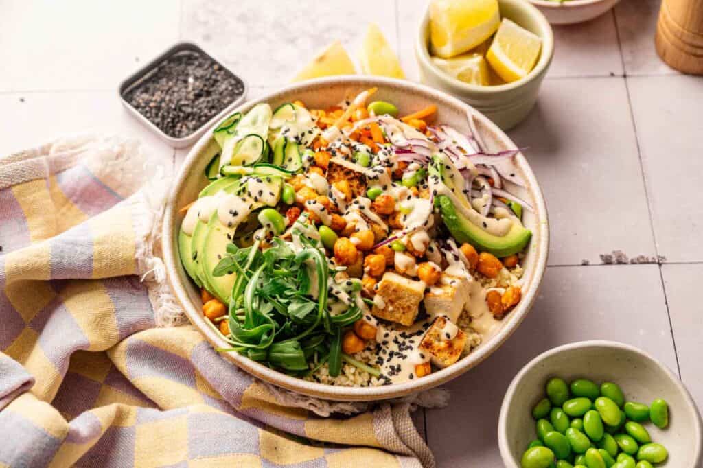 A vibrant Vegan Buddha Bowl filled with chickpeas, avocado, arugula, cucumber, shredded carrots, red onion, sesame seeds, and tofu in a creamy dressing, served alongside edamame and lemon wedges.