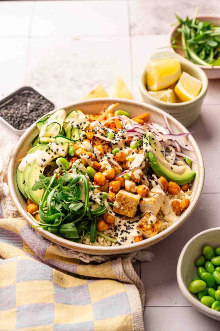A Vegan Buddha Bowl featuring tofu, chickpeas, avocado, greens, red onion, and black sesame seeds, all drizzled with creamy dressing. Served with lemon wedges and edamame on the side.