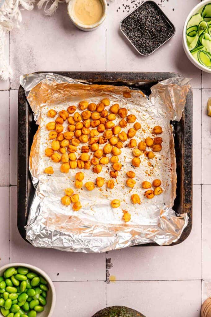 A baking tray lined with foil holds roasted chickpeas, surrounded by small bowls of edamame, sliced cucumber, black sesame seeds, and a creamy sauce.
