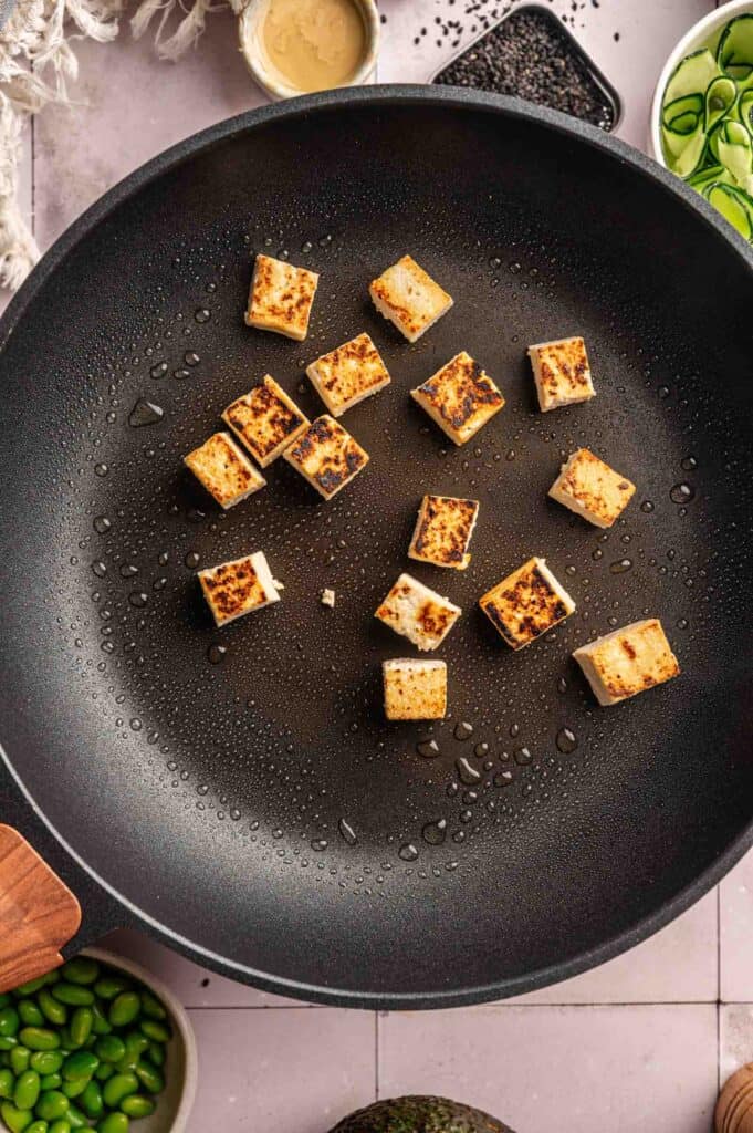 Cubed tofu pieces are browning in a black skillet, surrounded by small bowls containing sliced cucumber, edamame, and other ingredients—perfect for assembling a vibrant Vegan Buddha Bowl.
