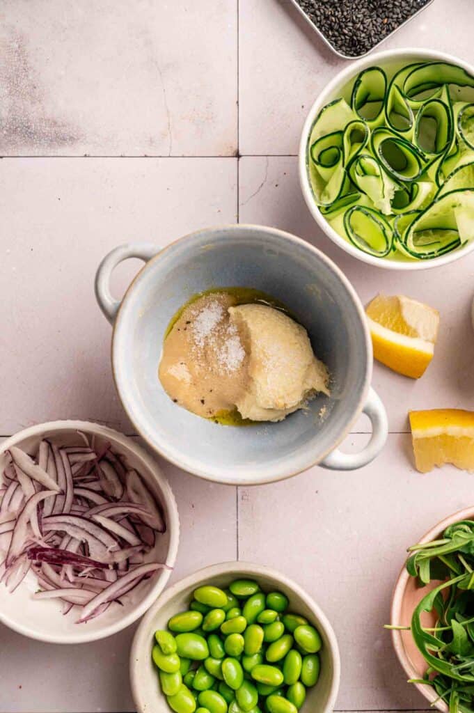 A Vegan Buddha Bowl takes center stage with a bowl of tahini, olive oil, and salt at its heart, surrounded by sliced red onions, edamame, cucumber ribbons, lemon wedges, arugula, and a bowl of black sesame seeds.