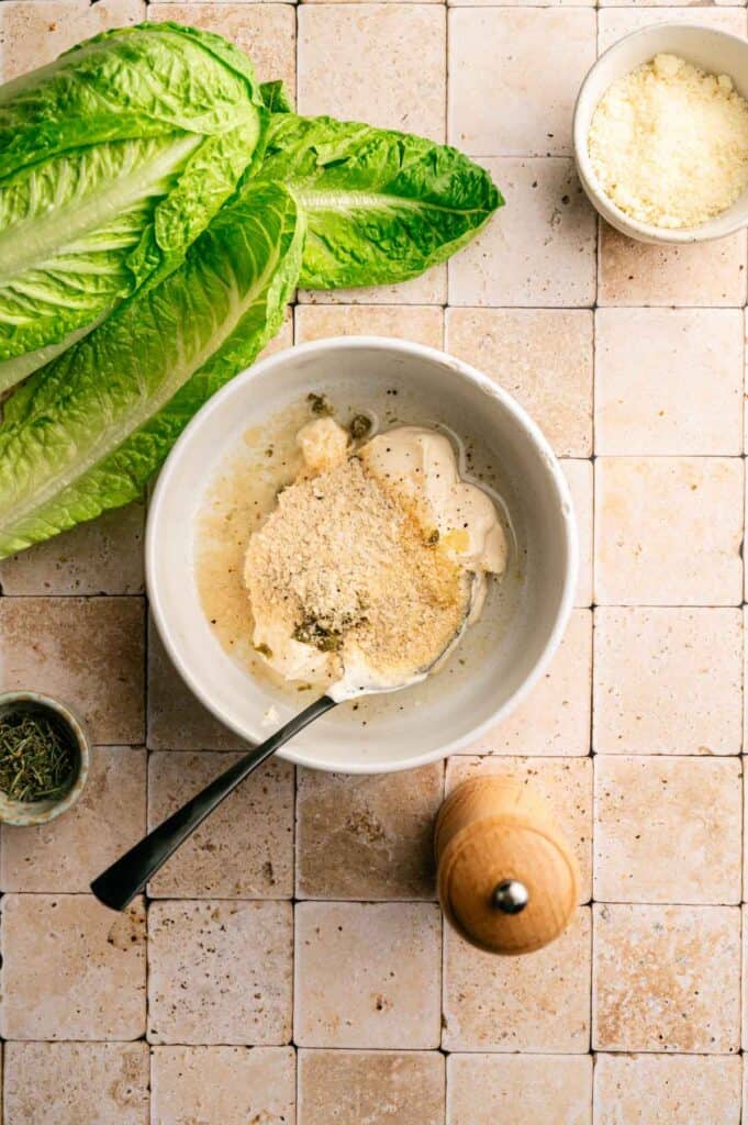 A bowl with Vegan Caesar Salad dressing ingredients, a spoon, romaine lettuce, grated cheese, dried herbs, and a pepper grinder on a tiled surface.