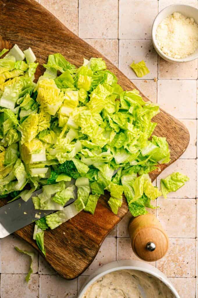 Chopped romaine lettuce on a wooden cutting board with a knife, next to a bowl of grated cheese and a pepper grinder on a tiled surface—everything you need to prepare a fresh Vegan Caesar Salad.