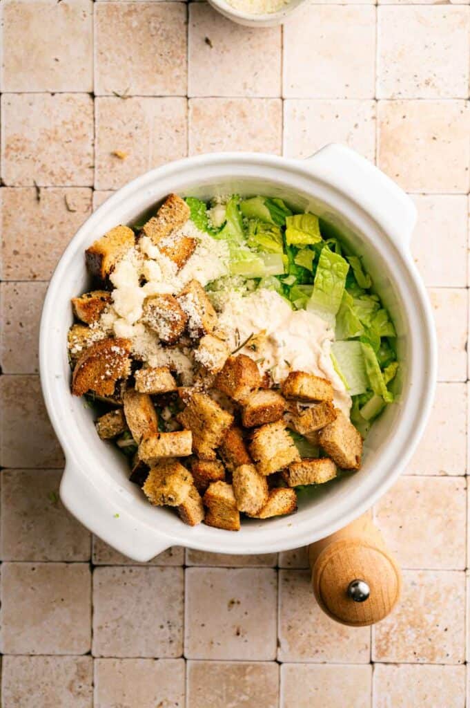 A white bowl contains chopped romaine lettuce, crunchy croutons, grated cheese, and creamy Vegan Caesar Salad dressing. A pepper grinder is next to the bowl on a tiled surface.