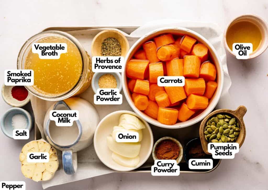 A tray with labeled ingredients for Air Fryer Vegan Carrot Soup: vegetable broth, smoked paprika, salt, garlic, coconut milk, herbs of Provence, garlic powder, onion, curry powder, carrots, cumin, pumpkin seeds, and olive oil.